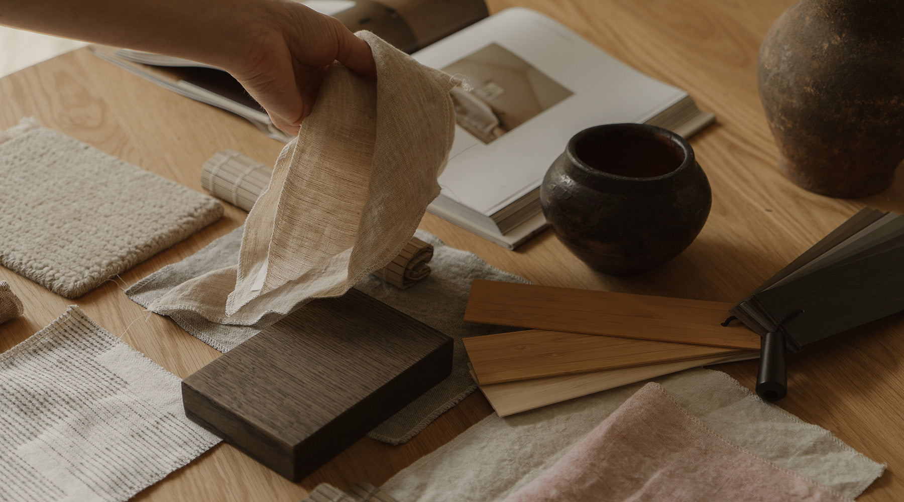 Person holding a fabric swatch over a wooden surface with color samples and a pot.