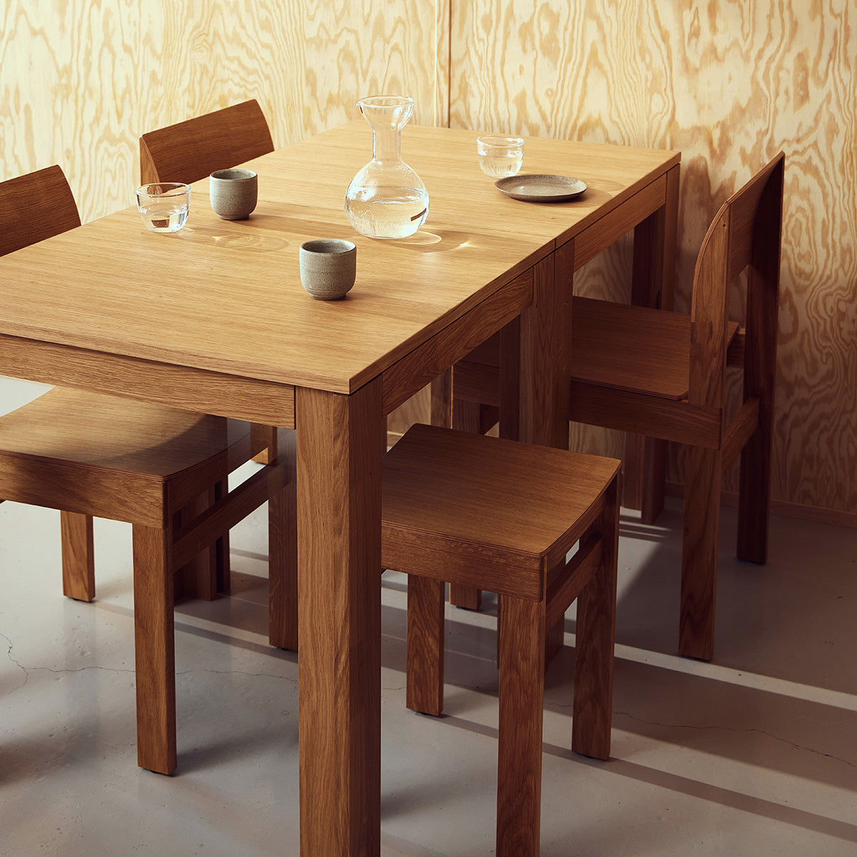 Vierkante tafel met ultra-dun tafelblad en verzonken poten van massief eikenhout. Subtiele schaduwlijn tussen blad en frame zorgt voor een licht zwevend effect. Modulair ontwerp dat meerdere tafels naadloos in een rij verbindt. Robuust en elegant, perfect voor dagelijks gebruik in moderne interieurs.