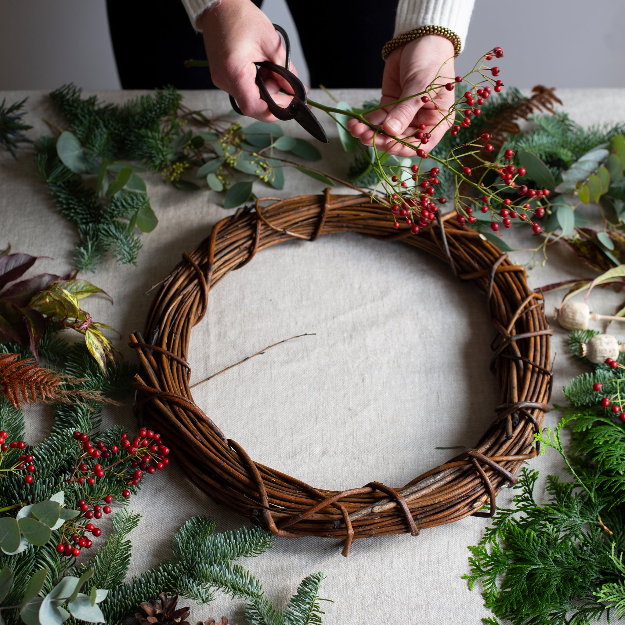 Person arranging greenery and red berries on a woven wreath on a light surface.