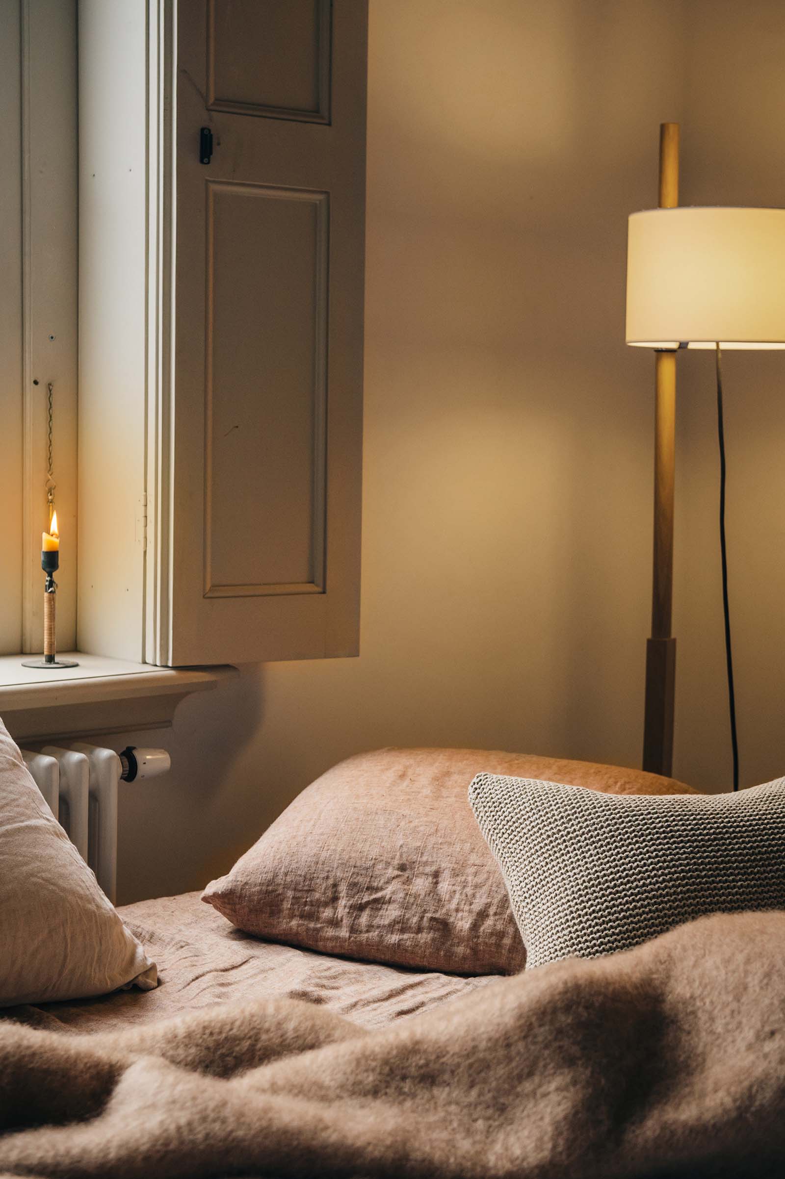 Hazel linnen kussens en mohair deken, op een bed voor een raam met vensters en een brandende kaars. In de hoek een TMM lamp van Santa & Cole.