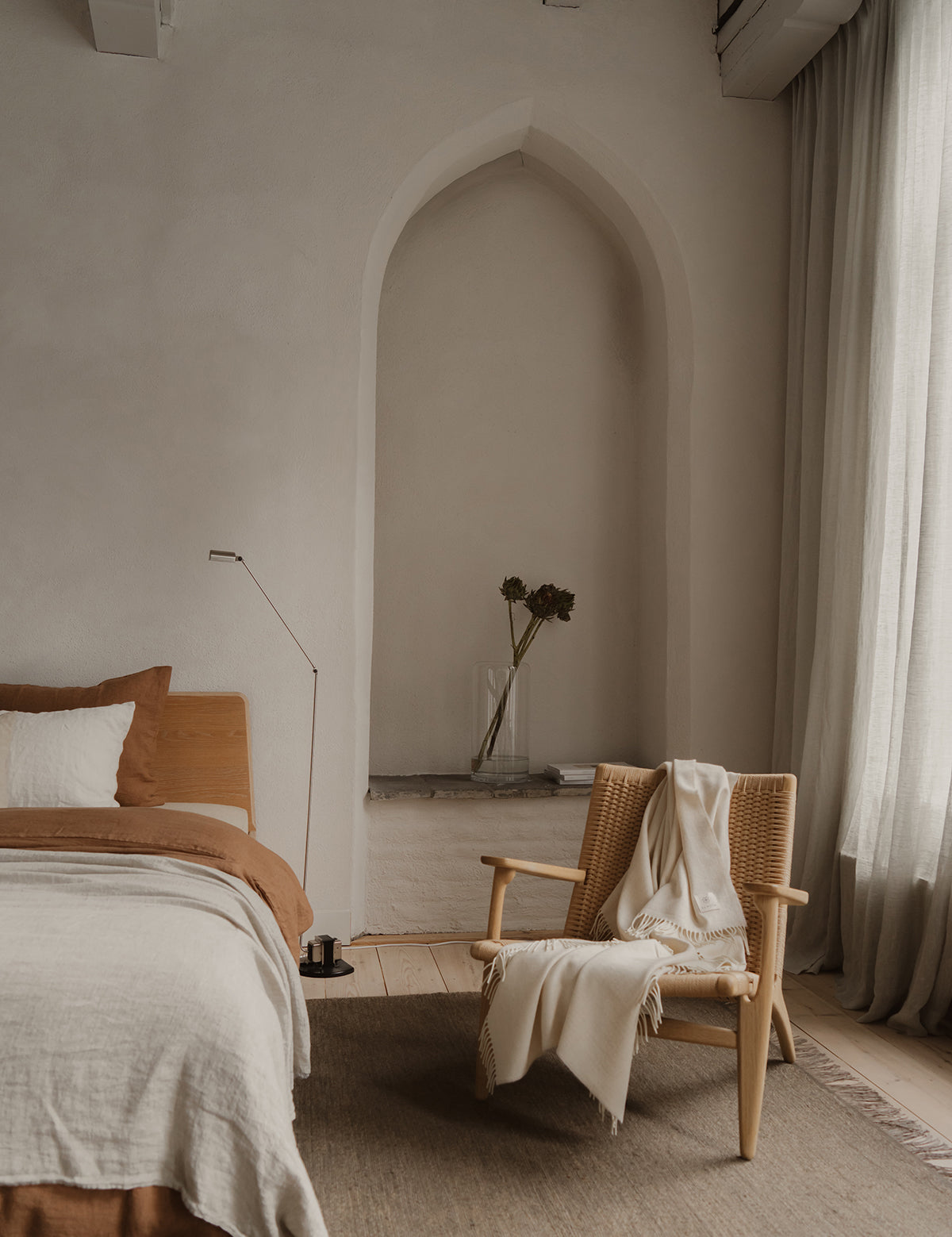 Slaapkamer met By Mölle inbetween linnen gordijnen en linnen beddengoed in cinnamon en flax. Op de houten vloer ligt een donker vloerkleed en er staat een lounge stoel met witte wollen deken.