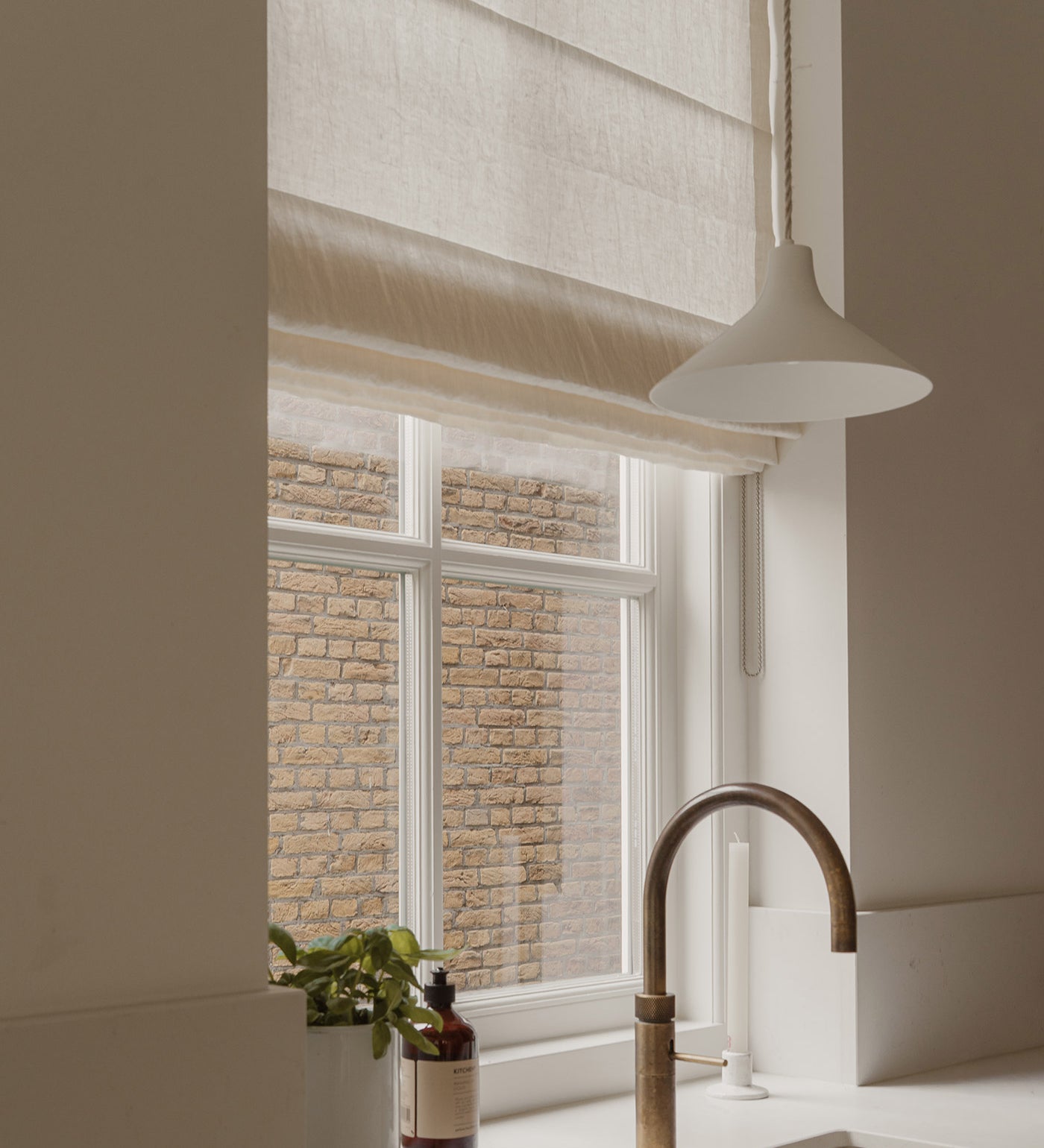 A modern kitchen scene featuring a beige linen Roman shade on a window, a white pendant light, a white sink, and a curved brass faucet, with a brick wall visible outside.