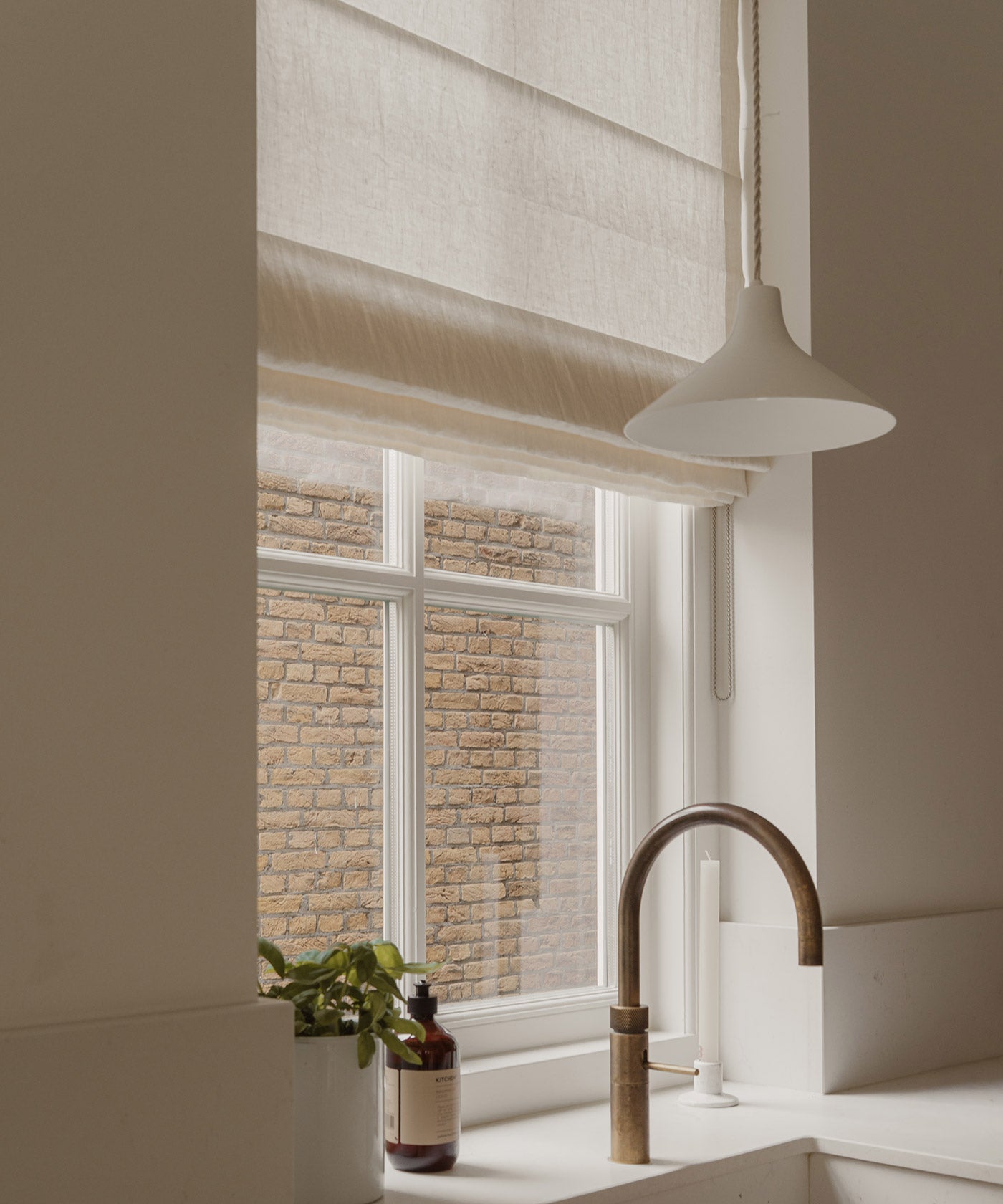 A modern kitchen scene featuring a beige linen Roman shade on a window, a white pendant light, a white sink, and a curved brass faucet, with a brick wall visible outside.