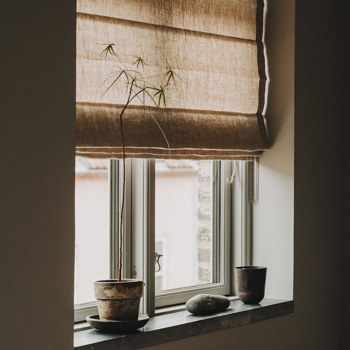 Puur linnen vouwgordijnen in de naturelle tint clay, in een raam met plant en decoratieve items.