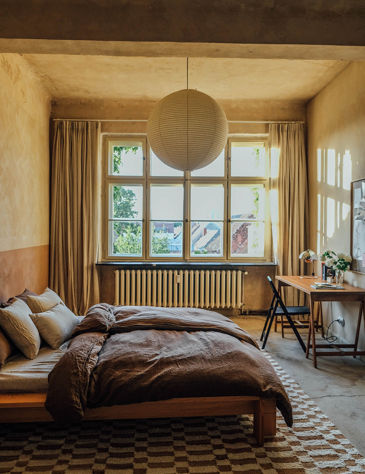 Gezellige slaapkamer in warme tinten, met puur linnen gordijnen en beddengoed van By Mölle, een bureau met bloemen en een geblokt vloerkleed.