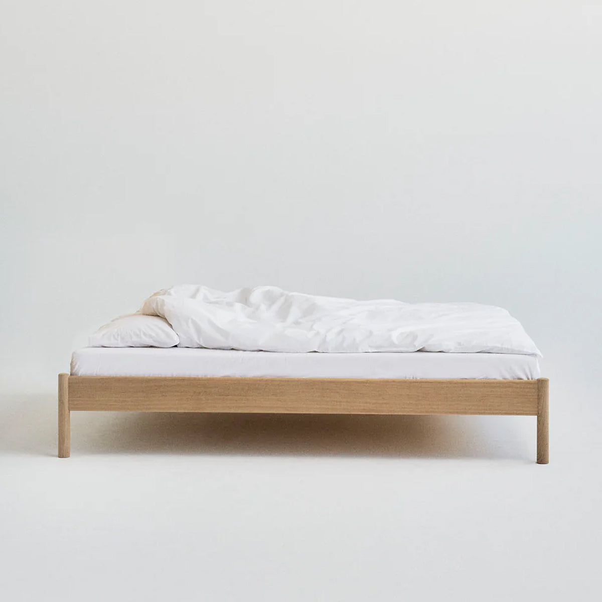 Minimalistisch houten bed Alken zonder hoofdbord, met wit beddengoed op een effen achtergrond.