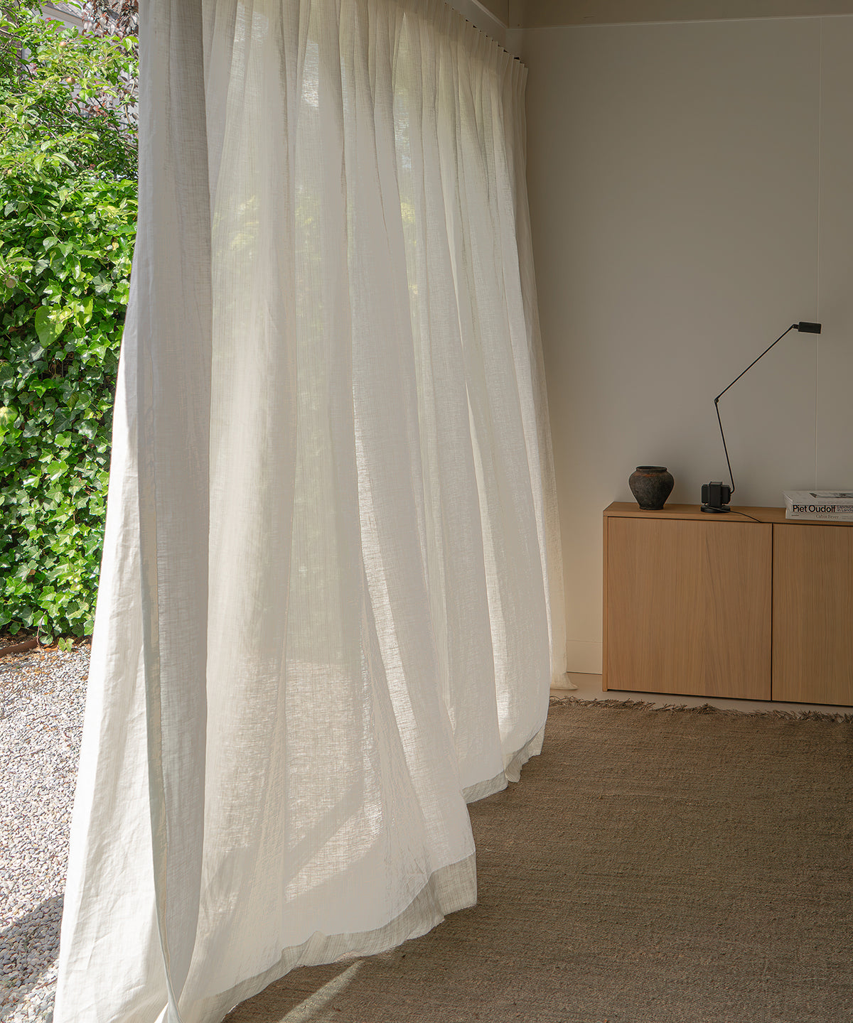 A long, semi-sheer white curtain panel hanging from a rod in a bright room, with a wooden cabinet, lamp, and outdoor greenery visible in the background.